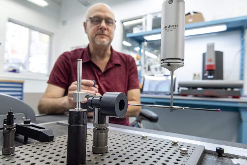 A man in a red shirt works in a laboratory, focusing on precision measurement instruments and equipment on a perforated metal table, with lab tools and machines in the background.