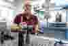 A man in a red shirt works in a laboratory, focusing on precision measurement instruments and equipment on a perforated metal table, with lab tools and machines in the background.