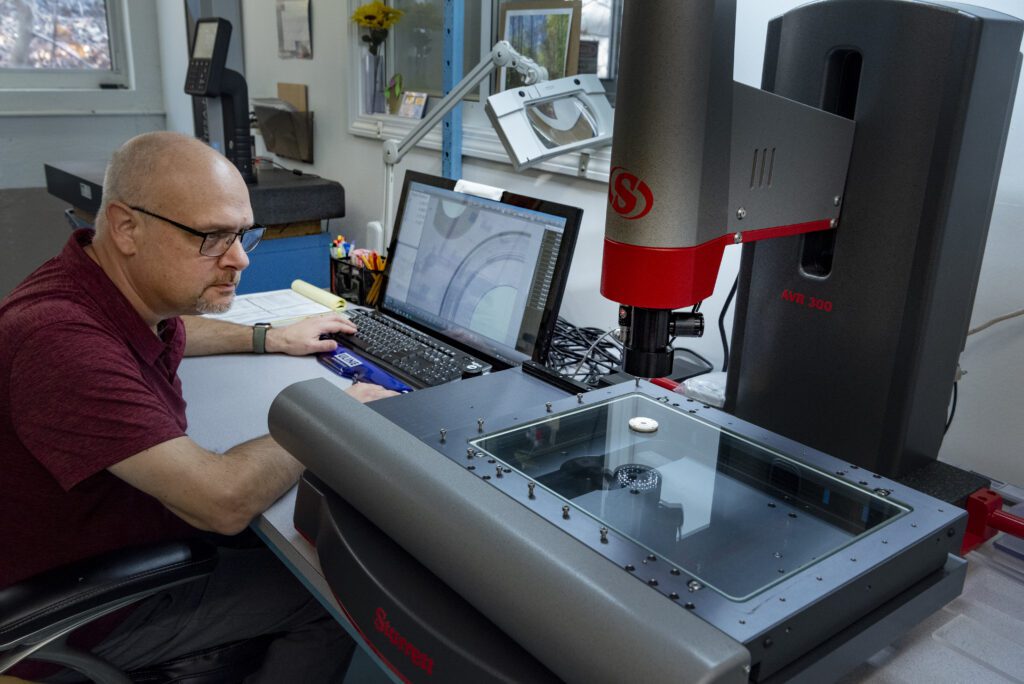 A man wearing glasses operates a laptop and monitors a measuring machine inspecting a plastic part in a laboratory or office setting. Various office supplies and tools are visible on the desk.