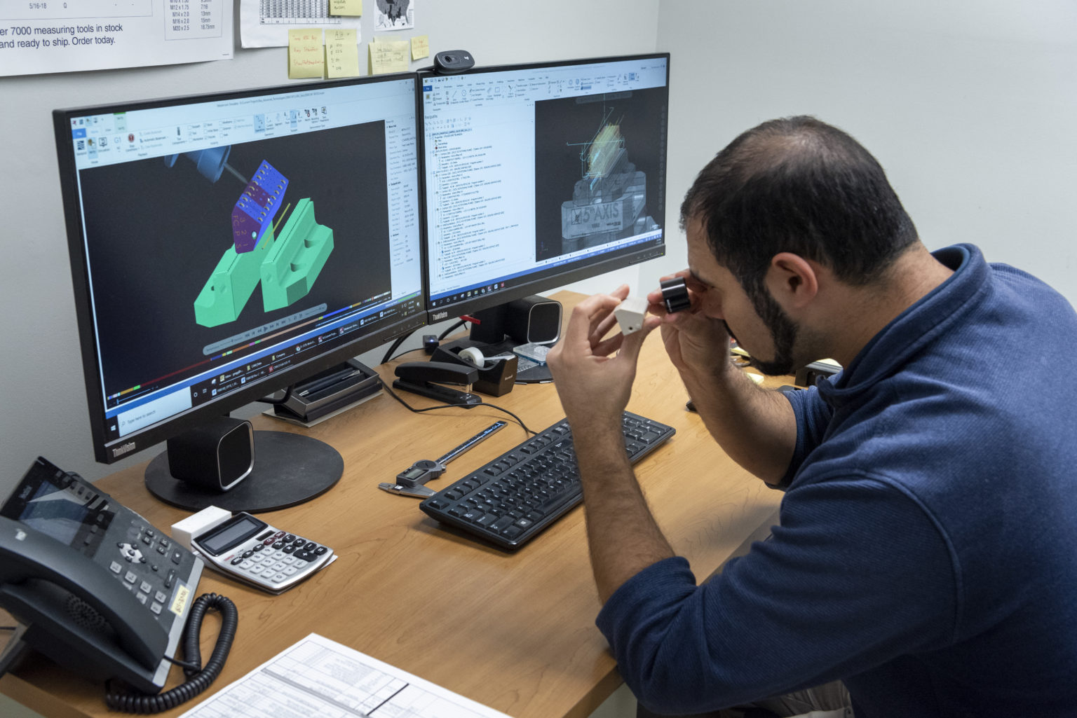 A man examines a small plastic component with a magnifier at his desk, which has a keyboard, phone, calculator, and paperwork. Two monitors display CAD models and technical drawings.