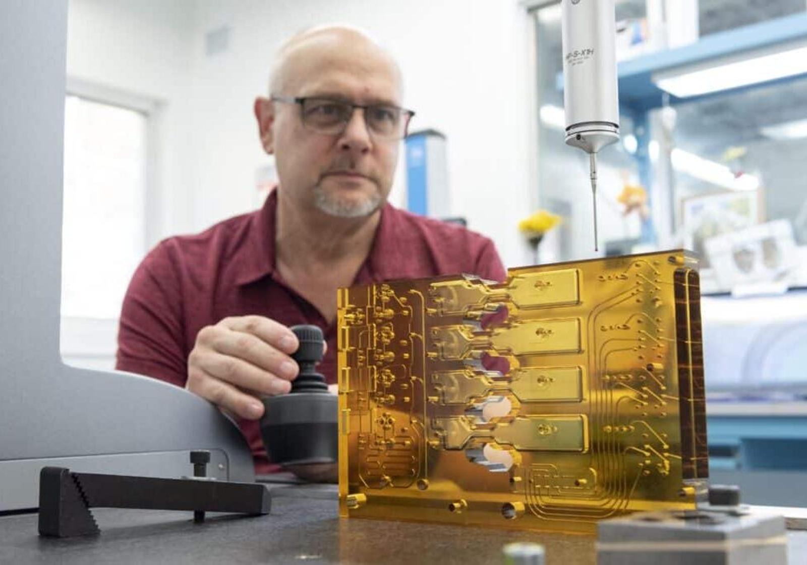 A man wearing glasses and a red shirt inspects a amber-colored ULTEM plastic manifold with a precision measuring device in a workshop setting.
