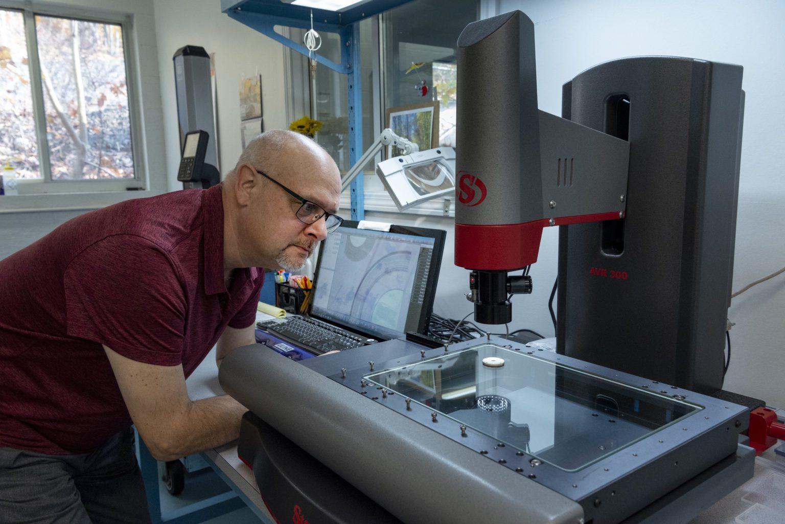 A man wearing glasses closely examines an object under a large digital microscope in a lab, with a laptop and various tools on the desk.