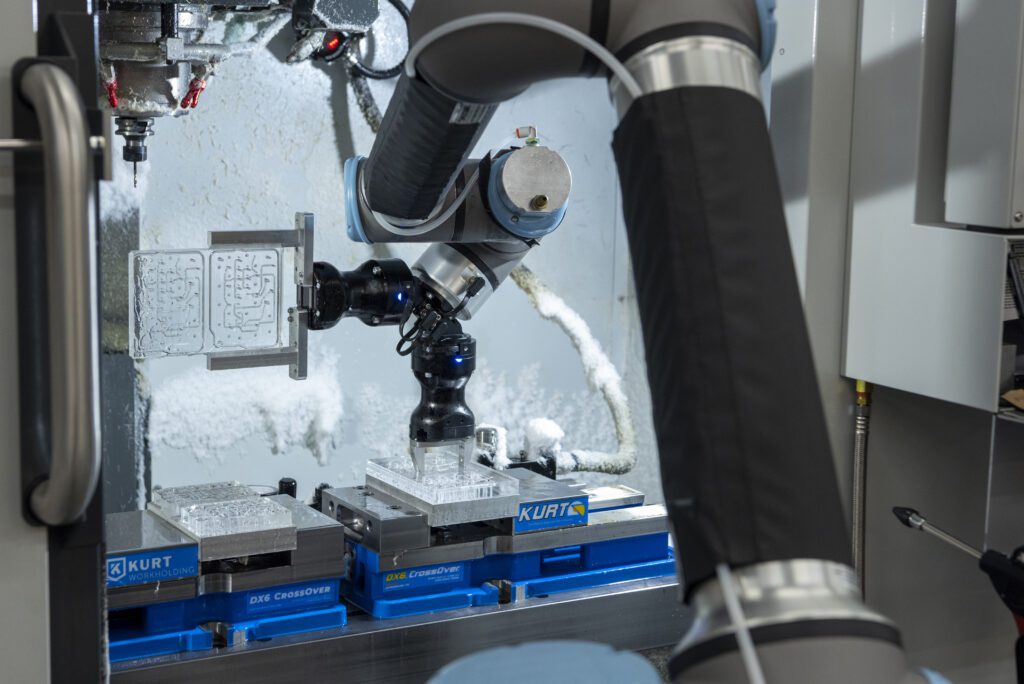 A robotic arm operates inside an industrial CNC machine, holding a tool above metal blocks secured on blue KURT vices, with a frosty, white substance visible on the surrounding surfaces.