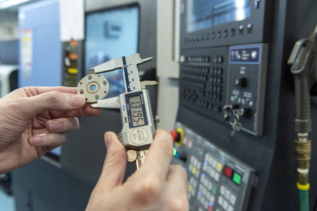 A person uses a digital caliper to measure a small PEEK plastic part in front of a control panel of a CNC machine. The caliper display shows a measurement.