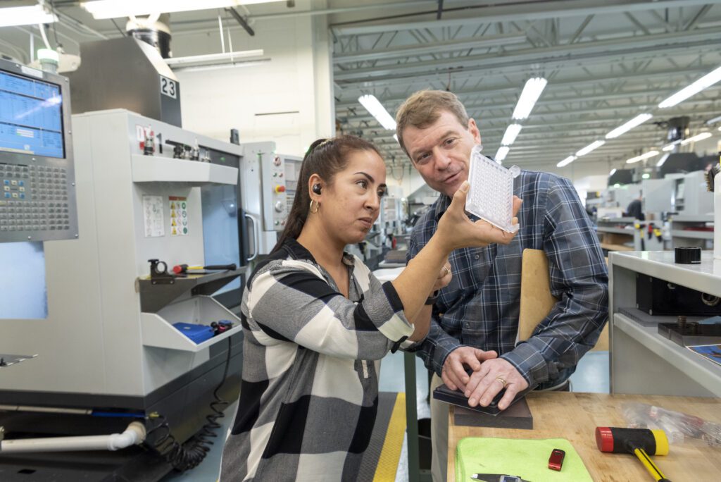 A woman shows a machined plastic manifold to a man in a factory. Both are wearing casual clothes and are discussing the manifold. Machinery and tools are visible in the background.