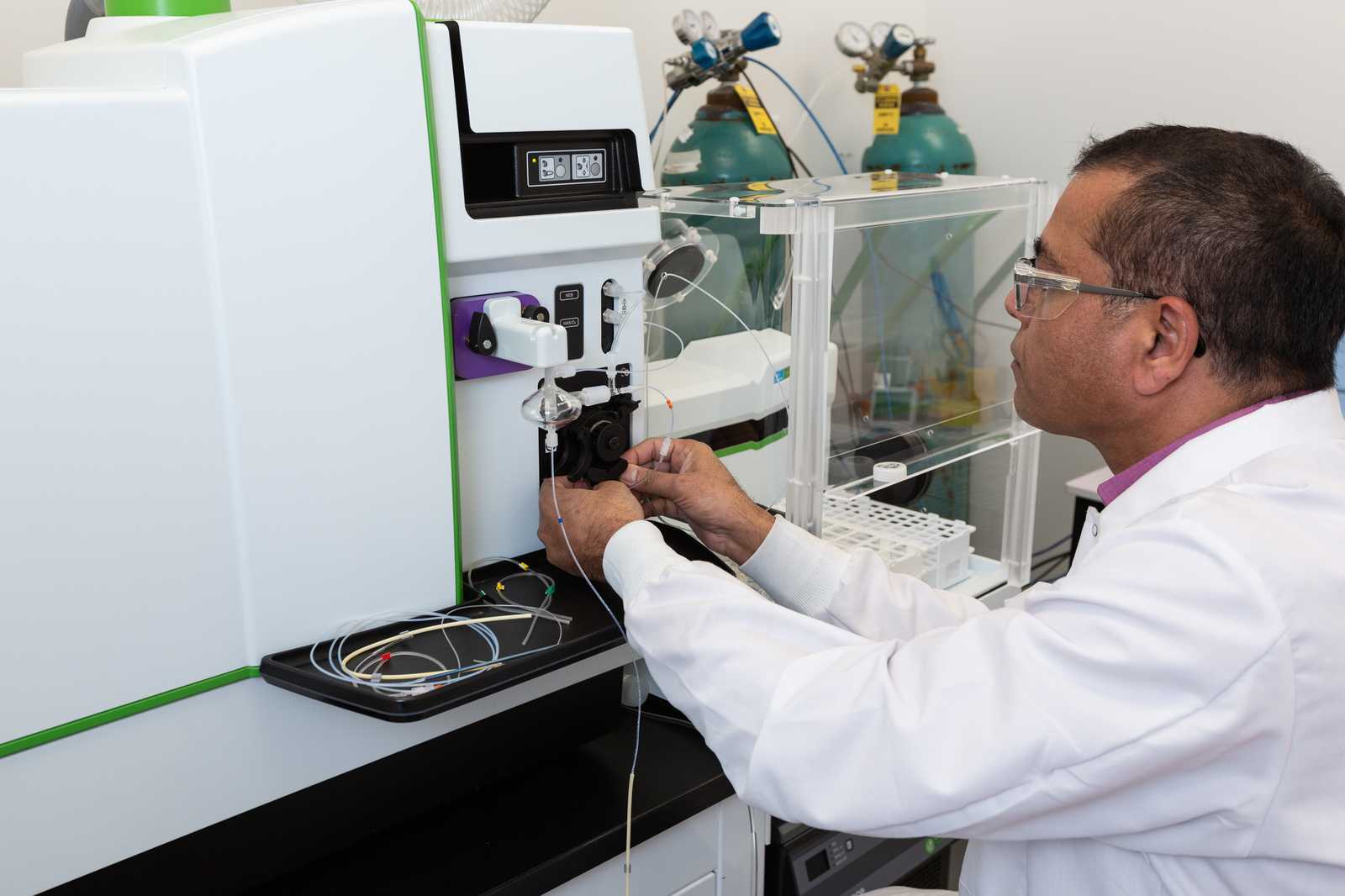 Person in a lab coat operating scientific equipment with gas cylinders and tubing in the background."