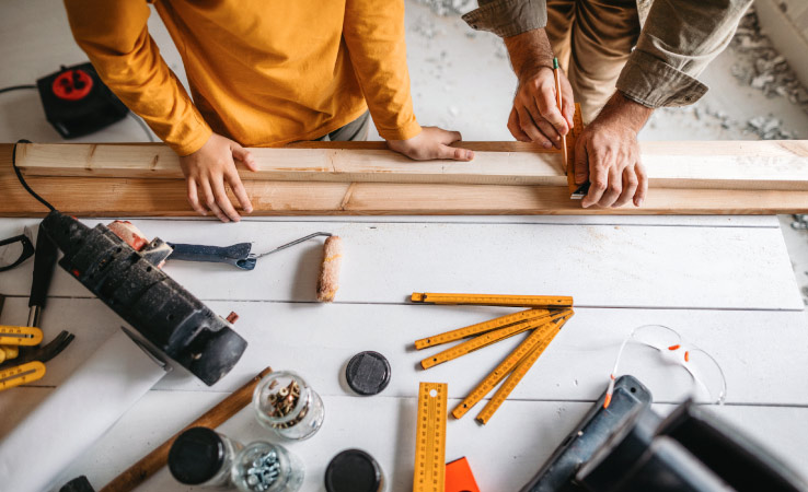 An aerial view of the hands of two men marking up a wood plank as part of a project. There is a white wood table with tools and rulers scattered.