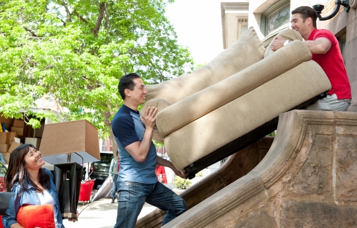 A woman is standing on a city sidewalk, smiling as two men carry a heavy, oversized chair up the steps to her new home. She’s hired local moving help to do the heavy lifting during her move.
