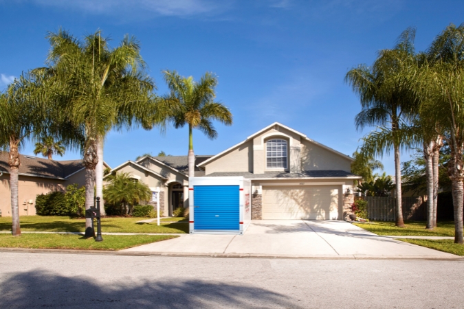 A portable storage container in Miami sits in the driveway of a two-story home with a manicured lawn and palm trees on either side.