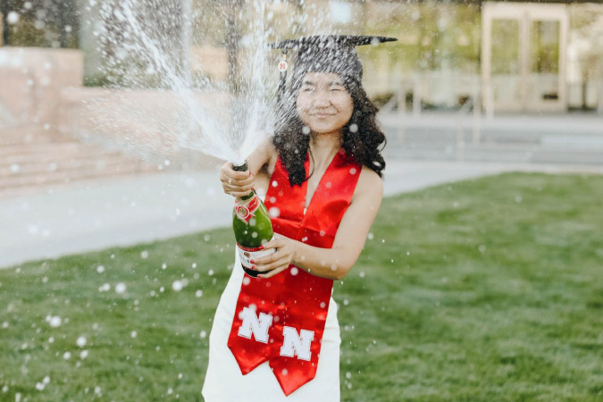 A young woman in a white dress with a black graduation cap and a red University of Nebraska Stole, opens a bottle of celebratory champagne to celebrate earning her degree.