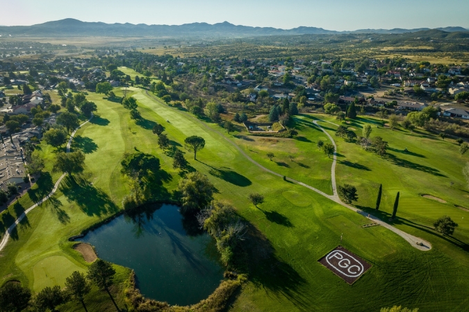 Aerial view of Prescott Golf Club featuring a pond, tree-lined fairways, a nearby neighborhood, and mountains in the background.