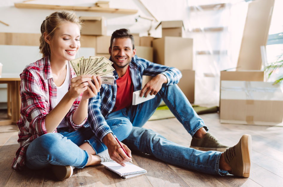 A couple is sitting on the floor surrounded by moving boxes. The woman is holding a stack of fanned-out bills, money they saved by using the cheapest way to move cross-country.