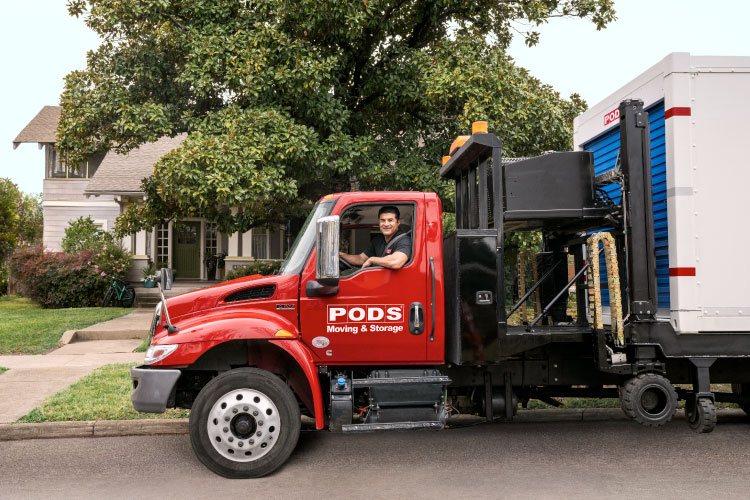 A PODS truck driver is smiling from the cab of his truck, parked in front of a residential home. There’s a PODS portable moving and storage container on the back of the truck. 