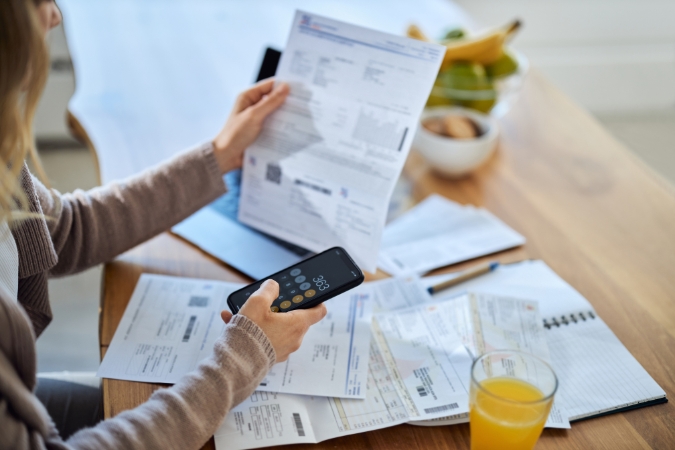 A woman is sitting at her kitchen table, using a calculator to add up household bills in an effort to answer the question “How much does a house cost?”