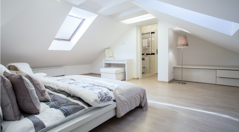 A modern bedroom with unique gabled ceilings and skylights. The walls are painted white, and the bed and other furniture are light-colored.