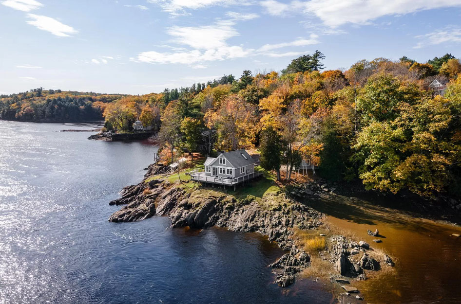 A waterfront home in Hampden, Maine, sits atop a rocky outcrop beside lush woodlands.