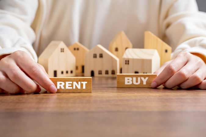 Close-up of a person sitting at a table with wooden model houses in front of them and two blocks with the words “RENT” and “BUY” printed on them.