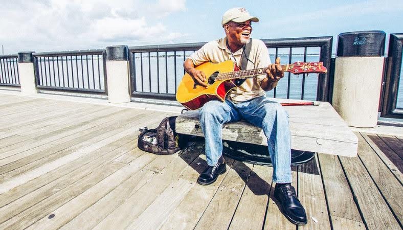 A local musician is sitting on a bench, singing and playing the guitar, on a boardwalk in Charleston, SC.