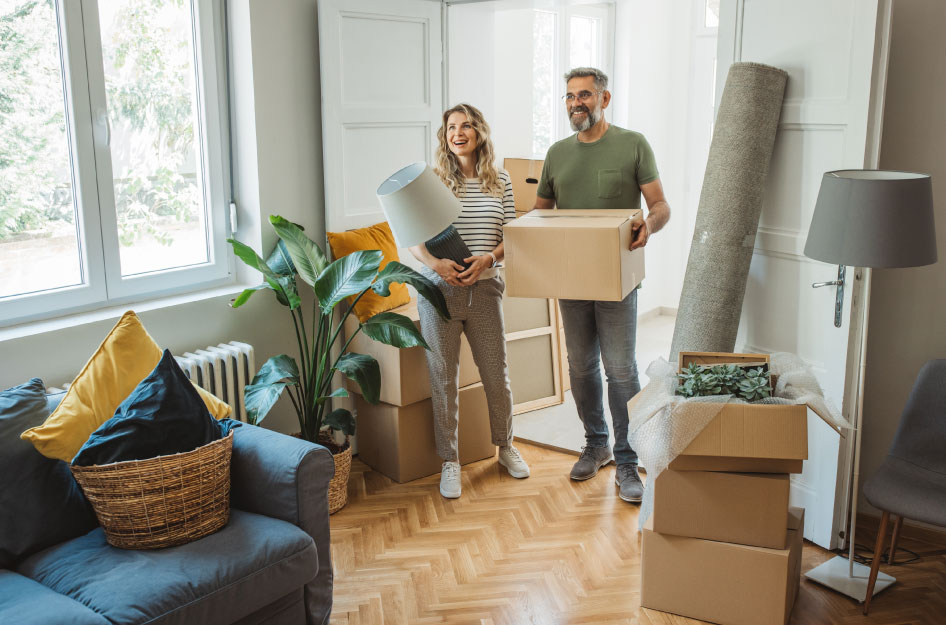 A happy couple is standing in the doorway of their new home with a moving box and a lamp, surrounded by furniture and other moving boxes, after using a complete moving timeline to organize their relocation.