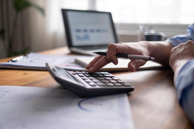 A person is sitting at a table, calculating the cost of moving using a calculator alongside various paper documents and a laptop.
