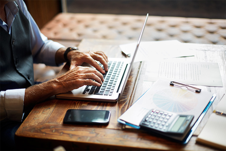 A retired man prepares his taxes on a laptop, with files and and a calculator close at hand