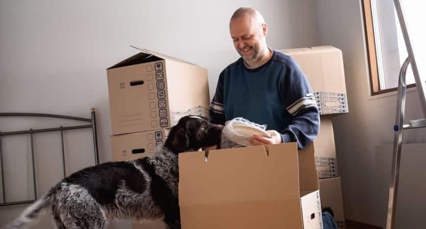 A man packing moving boxes with his dog nearby must consider his pet when deciding how to find a roomate.