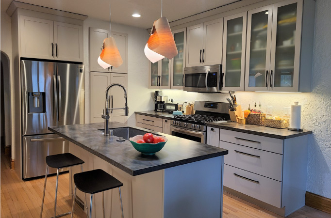 A newly remodeled kitchen with a combination of traditional cabinets and frosted window cabinets, all painted in a neutral gray. The kitchen also features a two-seater island with a built-in sink and new chrome appliances. 