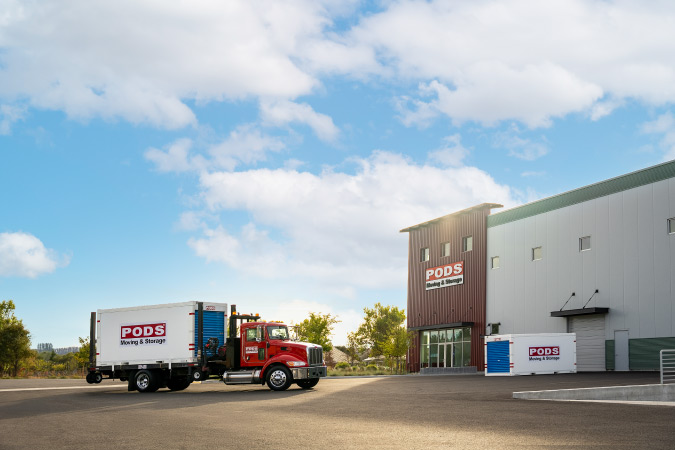 A PODS truck is parked in front of a secure PODS Storage Center. There is a PODS portable moving container on the back of the truck and another positioned beside the building.