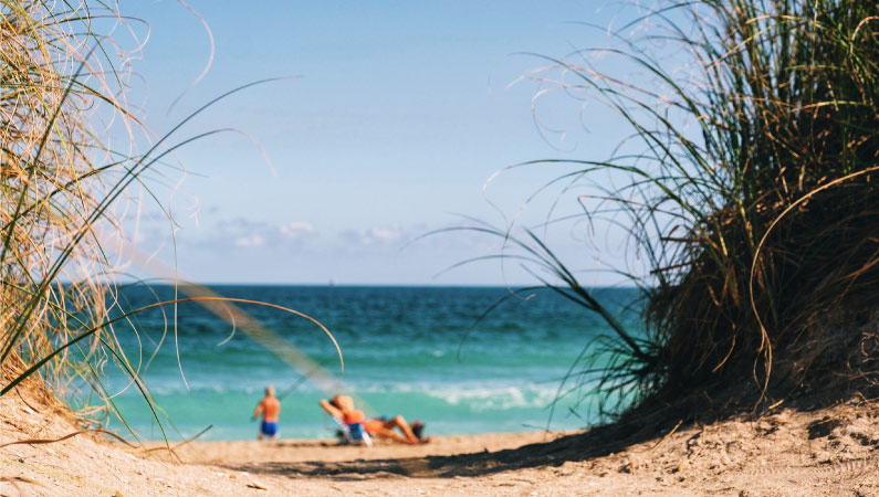 A retired couple enjoys a sunny day at a beach in Florida. The man is casting his fishing line into the clear, blue waters while his wife is sunbathing nearby.