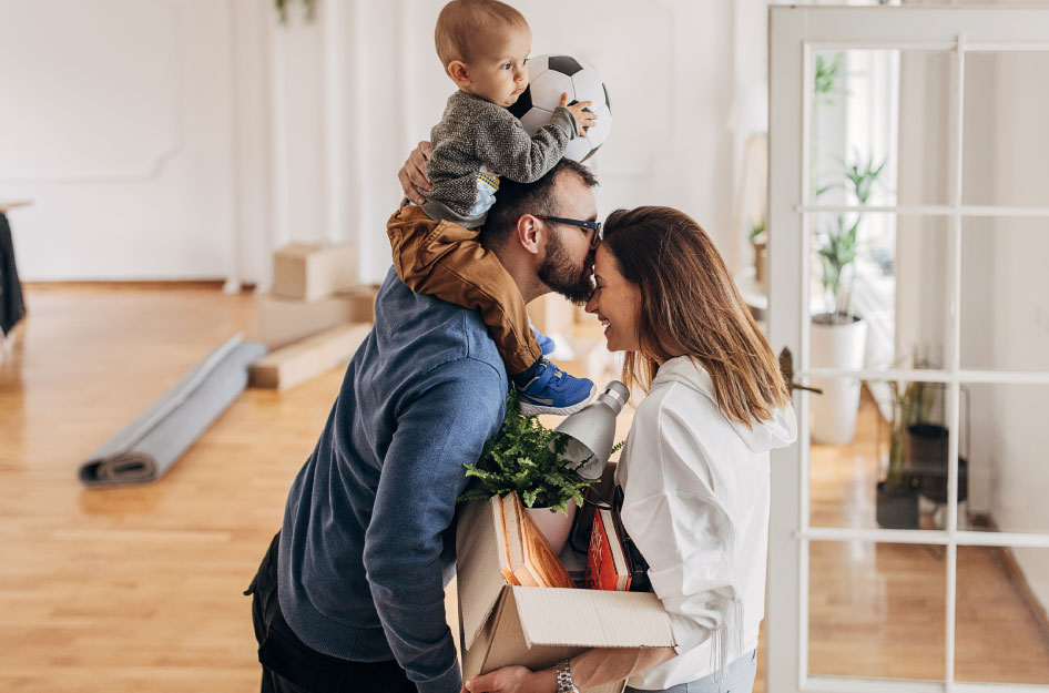 A couple preparing for moving with a baby playfully embraces with a half-filled moving box between them and a baby on the dad’s shoulders.