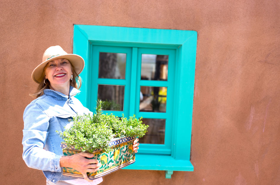 A senior woman is smiling in front of a classic New Mexico adobe house, holding an ornate flower box.