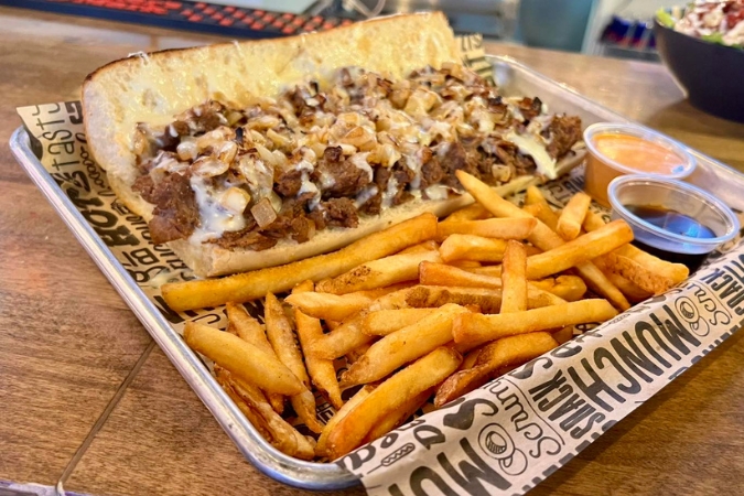 A philly cheesesteak sandwich and french fries with sauces on the side on a serving plate from Townies Philly Kitchen and Bar, one of the best restaurants in Rochester, MN.