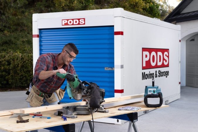 A man is cutting a plank of wood at a sawhorse in front of a PODS portable storage container, which he’s using for storage during home renovation.