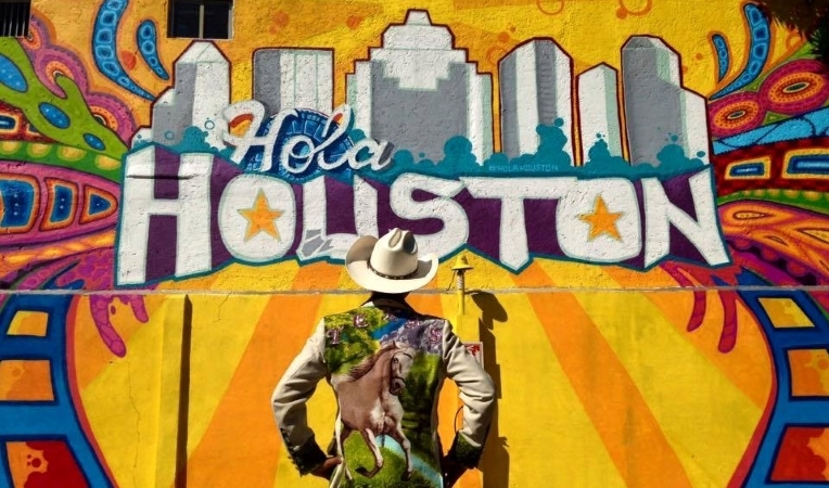 A cowboy in a white hat and painted blazer is standing in front of a colorful mural that reads, “Hola Houston.”