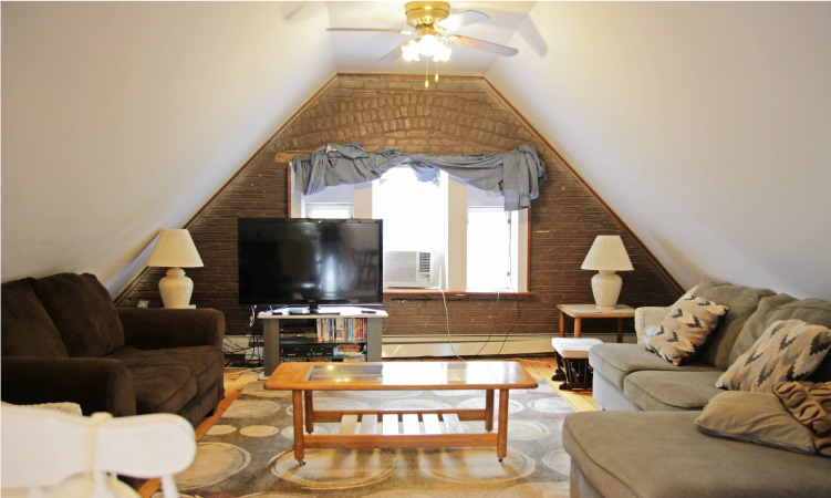 An attic that has been renovated into a cozy living room space with comfortable furniture and a TV. A large window is letting natural light into the room.