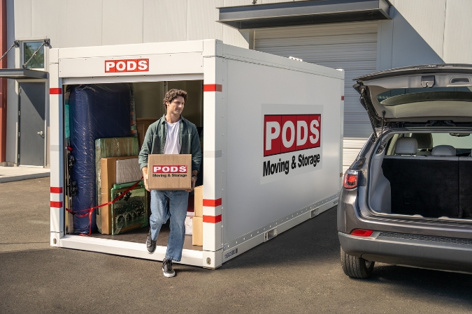 A young man is in the process of moving home after college, carrying moving boxes from a PODS portable moving container to the trunk of his car.
