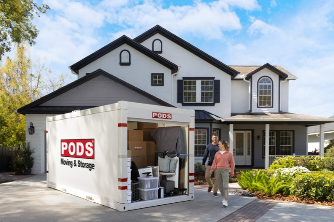 A couple is walking toward a loaded PODS on-site storage container, which is sitting in the driveway of their two-story home.