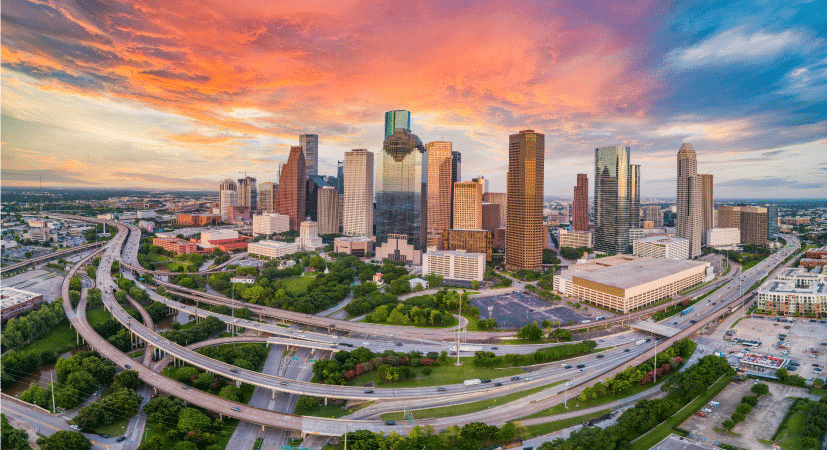 Aerial view of Houston, Texas, during a vibrant sunrise, featuring half a dozen curving freeways wrapped around Downtown Houston with lush greenery filling the gaps between them.