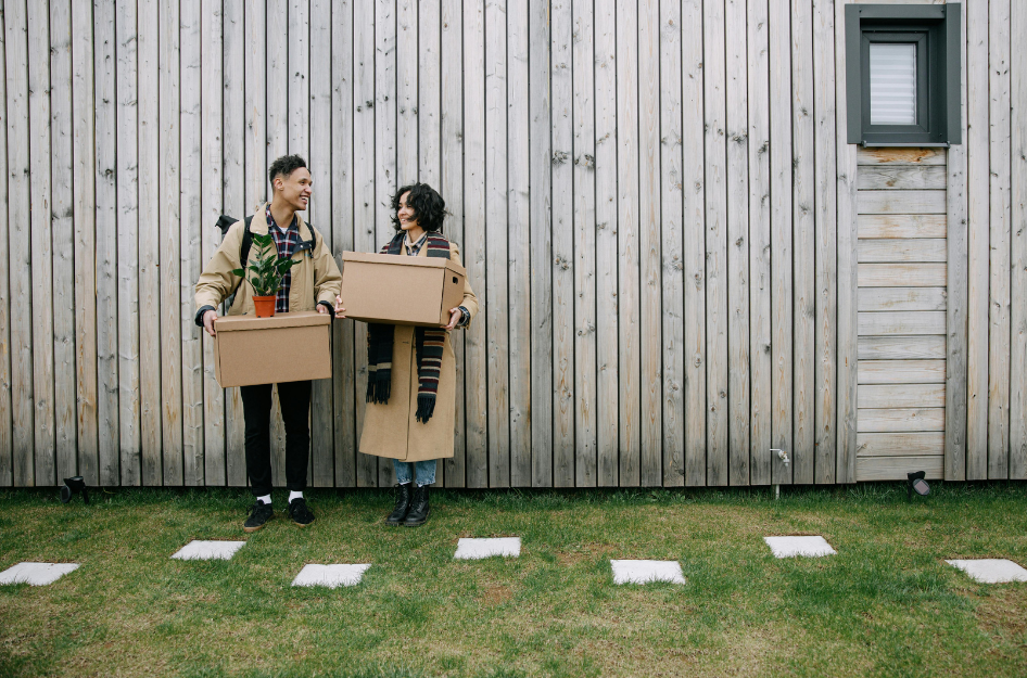 A young couple stands outside their new home holding moving boxes and a house plant.