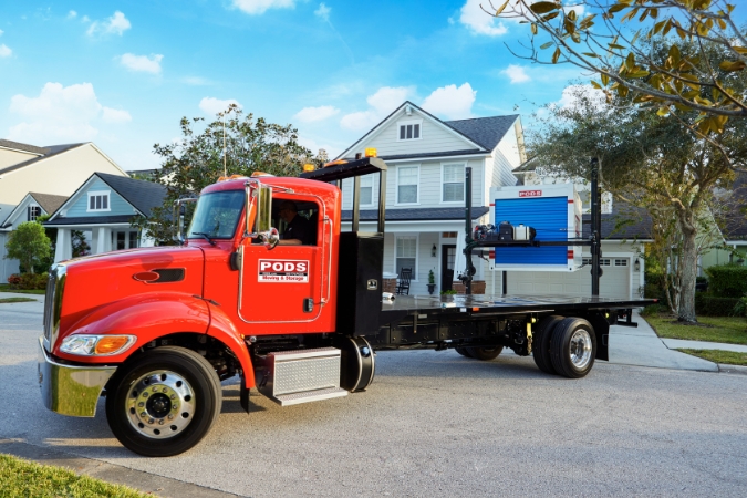 A PODS truck is parked outside a residential home as a portable storage and moving container is being positioned in the driveway with the help of PODZILLA.