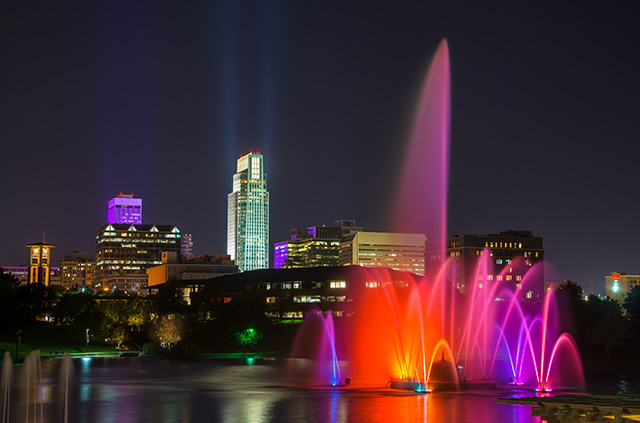 The Omaha skyline at night. A fountain is bathed in red and purple, and the buildings are lit in shades of green, white, and purple.