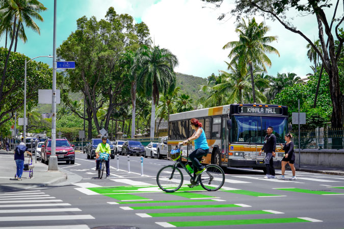 View from across the street of a Honolulu city bus waiting at a stop as pedestrians and a bicyclist cross in front.