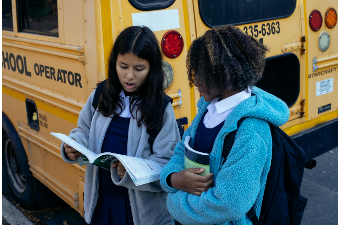 Two young girls stand in front of a school bus reading a book.