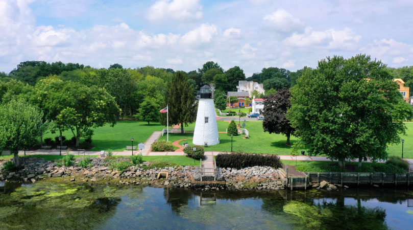 A view of Concord Point Lighthouse in Havre de Grace, Maryland, from the water. The lighthouse appears a bit stout. Its white tower is wider at the base, gradually becoming more narrow until it reaches the black lantern room. The photo was taken on a beautiful summer day. The lawn and surrounding trees are lush and green and the sky is a light blue, filled with fluffy, white clouds. 