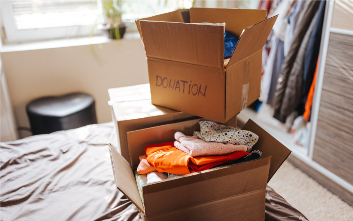 Three cardboard boxes are stacked on a bed. The boxes are labeled “DONATION” and are filled with clothing to be donated.