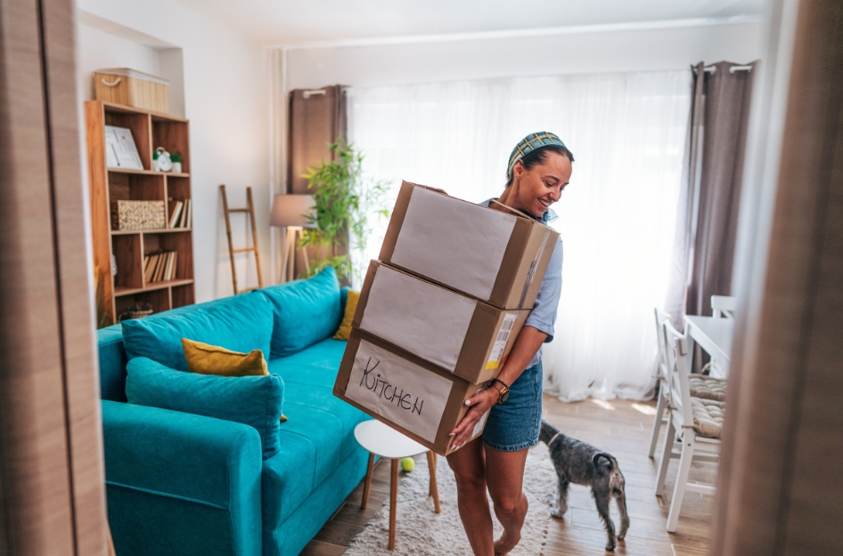 A young woman is carrying a stack of moving boxes past her small dog and through her living room as she prepares for a long-distance move.