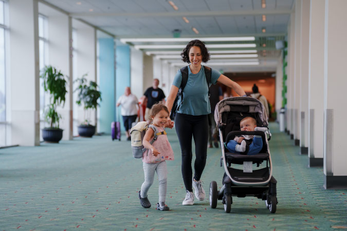 A mom moving long-distance with baby and toddler walks through an airport holding her toddler’s hand with one hand and pushing a stroller with the other.