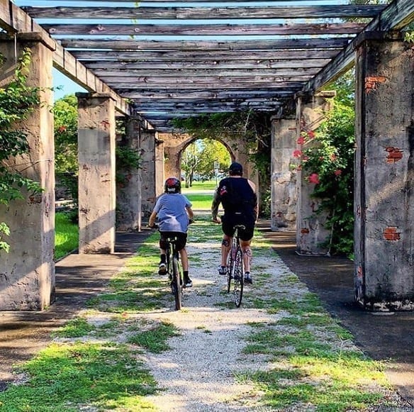 A pair of bikers bike down a path covered by a building in the town of Coral Gables. 