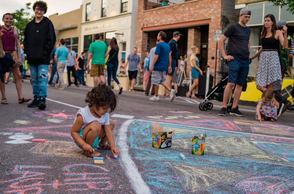 A young kid is drawing with chalk, surrounded by family and friends at a summer block party in one of the best neighborhoods in Denver for families.