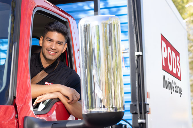 A PODS driver is smiling out the driver’s side window of the parked truck and there is a PODS portable storage container on the back of the truck.
